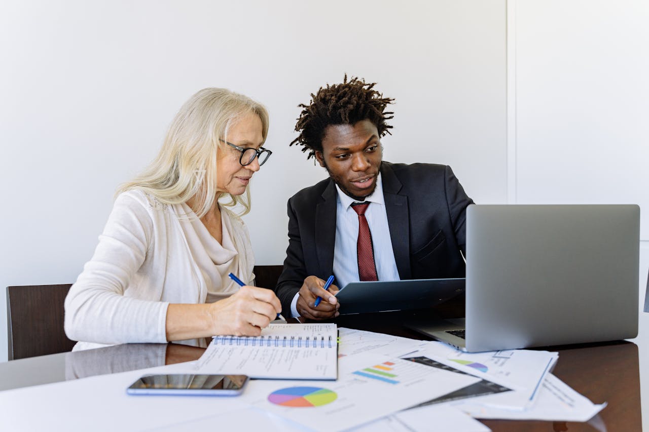 Two business professionals collaborating at a desk with laptops and documents in an office setting.